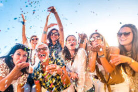 Cheerful group of happy people mixed ages generations women having fun all together during celebration party or carnival - view of fiends blowing. coloured confetti and laugh a lot in friendship